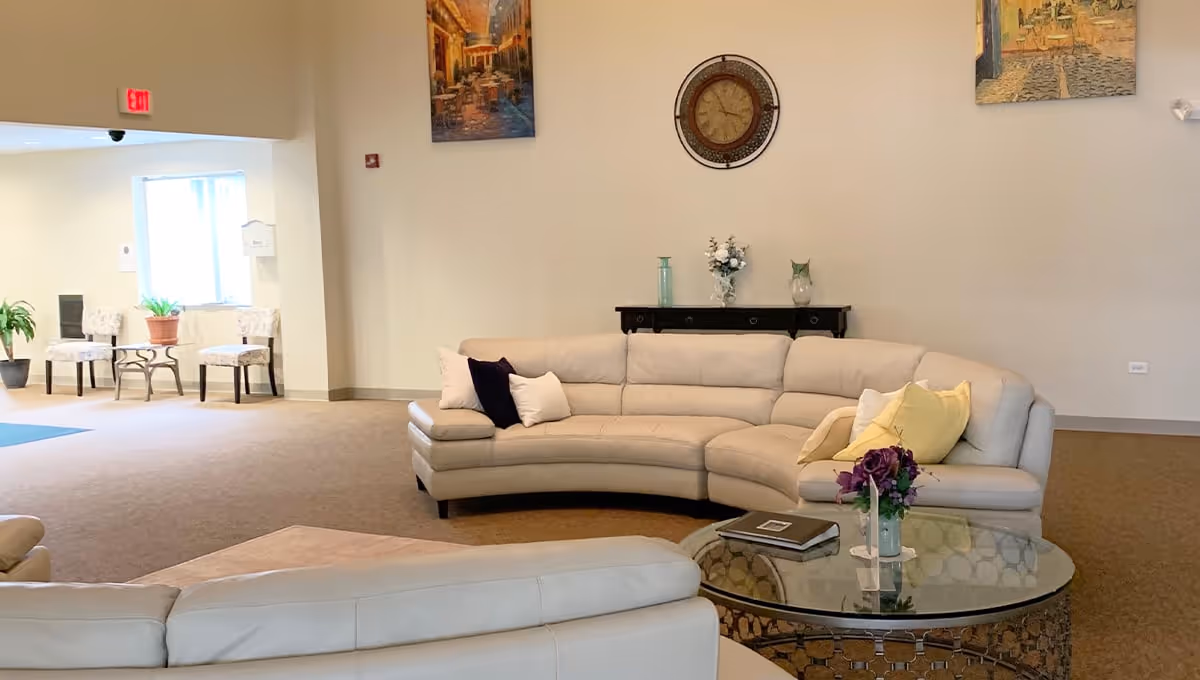 A spacious living room area in Poplar Creek Village featuring a large curved beige sectional sofa with multiple throw pillows, a round glass coffee table with a flower vase and a book, a dark wooden console table with decorative items, a wall clock, and two framed paintings on the wall. There are also two chairs and a small table near a window in the background.