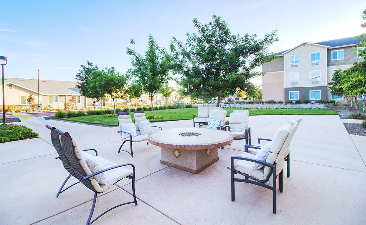 Outdoor seating area with cushioned chairs arranged around a circular fire pit on a concrete patio, surrounded by green grass, trees, and buildings in the background under a clear blue sky.