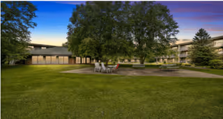 A spacious outdoor courtyard area at Heathers Estates featuring green grass, large trees, and several seating areas with tables and chairs under a clear sky at dusk.