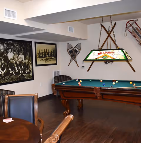 A recreational room featuring a billiards table with balls arranged on it, a hanging stained glass lamp with the word 'BILLIARDS', vintage snowshoes and skis mounted on the wall, two framed black and white photographs, and a round wooden table with chairs in the foreground.