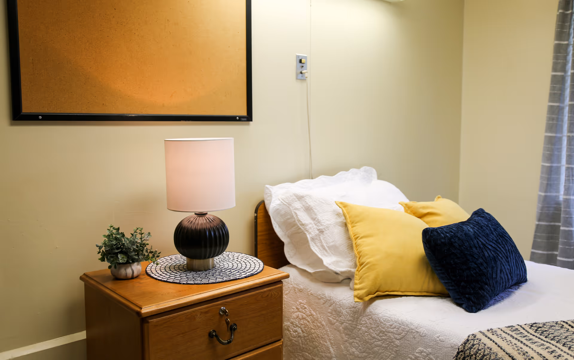 A cozy bedroom corner with a wooden nightstand holding a black lamp with a white shade and a small potted plant. Next to the nightstand is a bed with white bedding and three decorative pillows in yellow and dark blue. A window with gray curtains is partially visible on the right side.