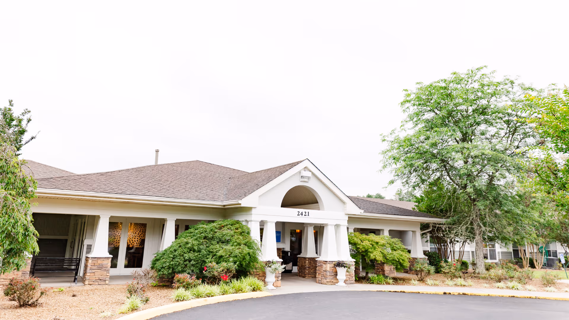 Front exterior view of a single-story senior living facility building with a covered entrance supported by white columns and stone bases. The building has a brown shingled roof and is surrounded by landscaped bushes, small trees, and plants. The address number 2421 is visible above the entrance.