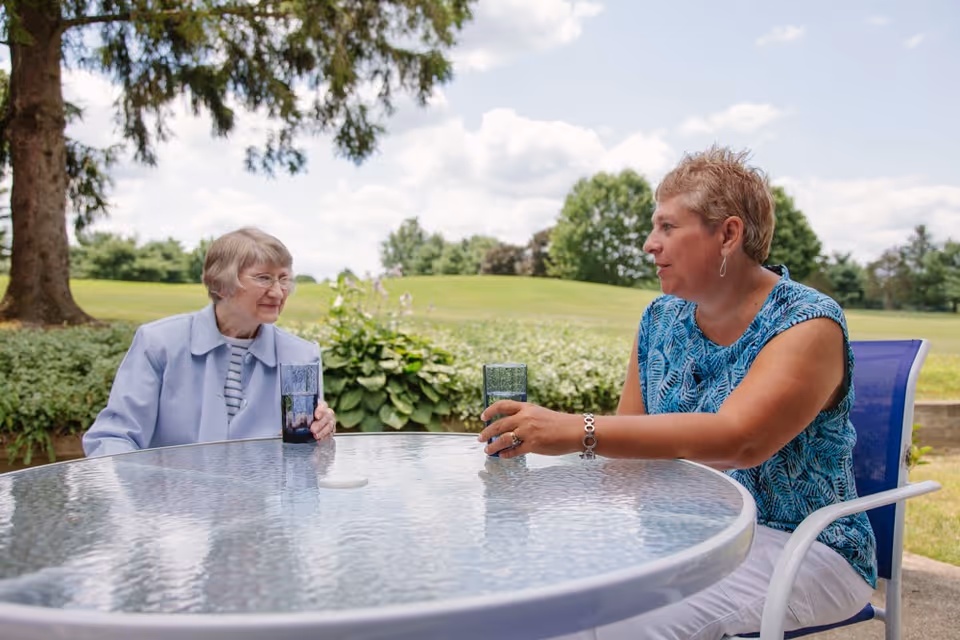 Two women sitting at a round glass outdoor table, each holding a glass of water, with greenery and trees in the background under a partly cloudy sky.