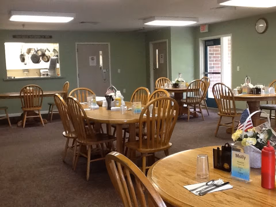 Interior view of a dining room in a residential care facility with several round wooden tables and chairs. Each table has condiments, utensils, and small decorative items. There is a serving window to the kitchen on the left wall and a door leading outside on the right. The walls are painted green and the floor is carpeted.