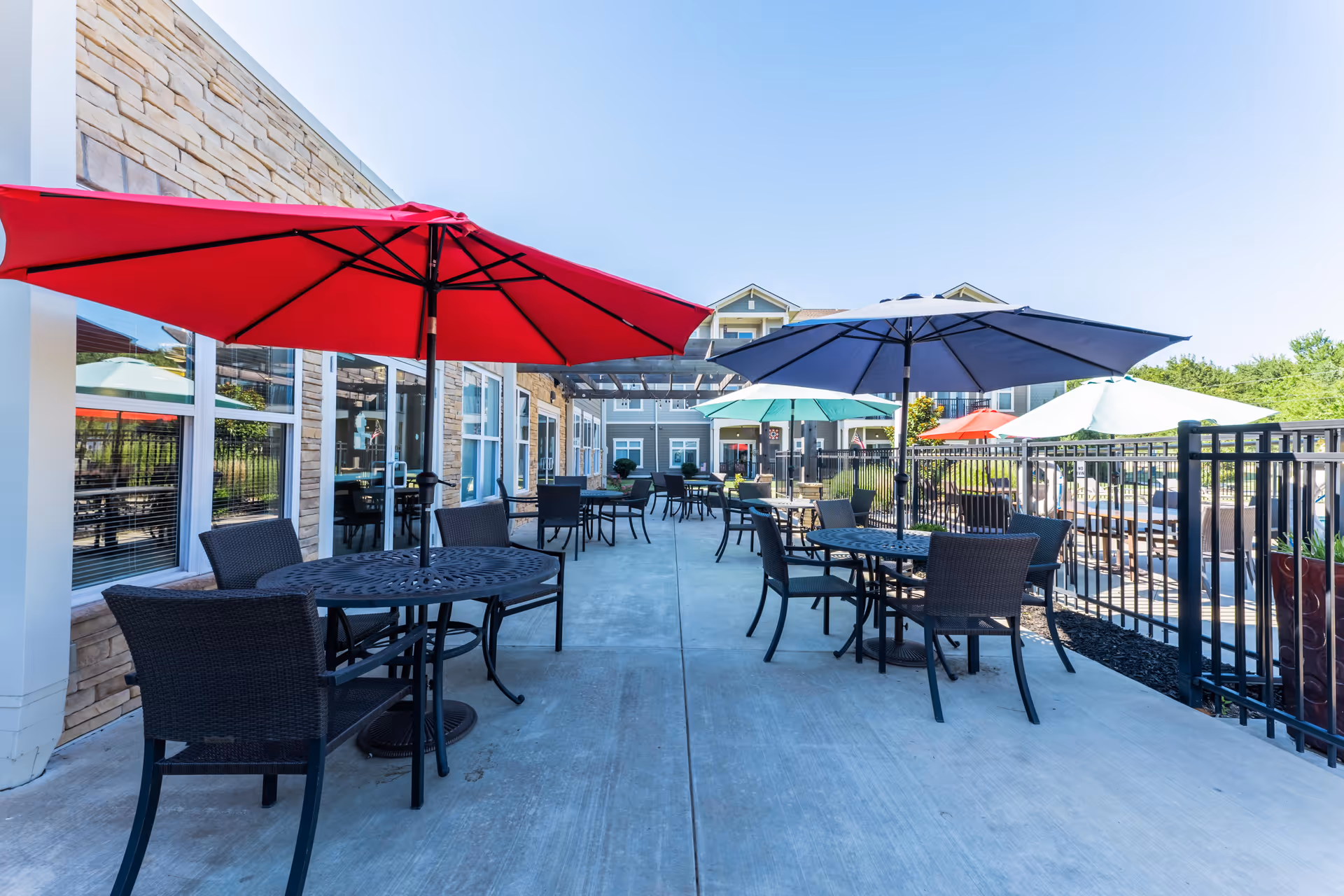 Outdoor patio area at The Cedars at Woodland Hills with multiple round tables and chairs under colorful umbrellas including red, blue, and green. The patio is paved with concrete and is adjacent to a building with large windows and stone accents. A black metal fence runs along one side of the patio.