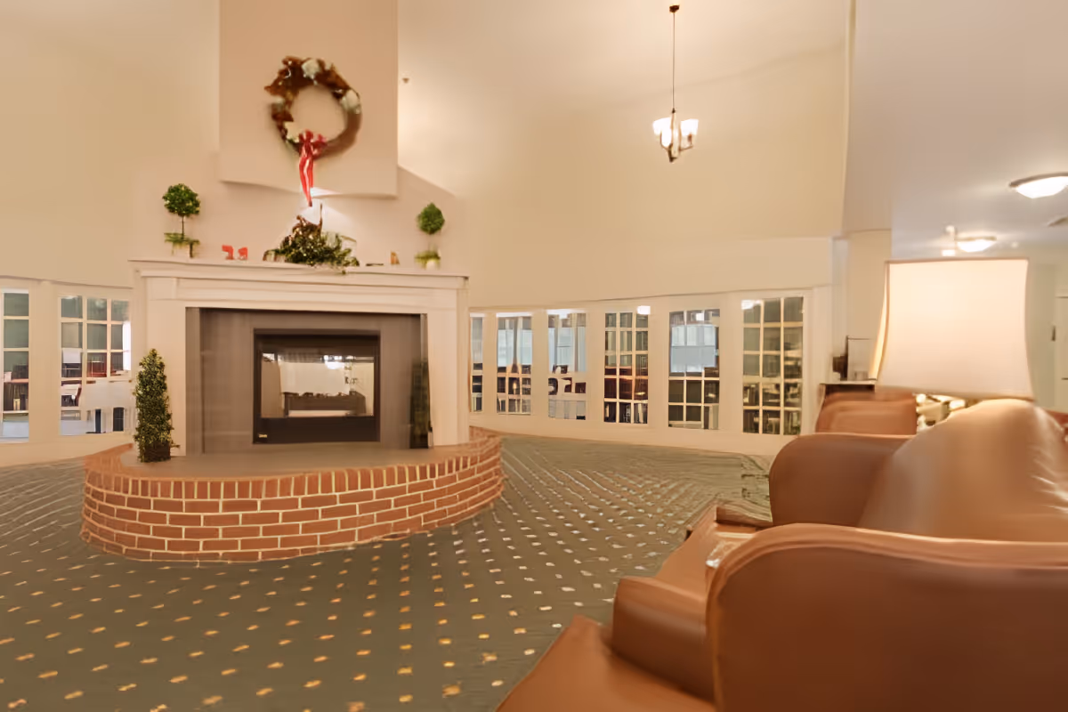 Spacious lounge with a central brick-surrounded fireplace, wreath above, and upholstered chairs under a high ceiling.