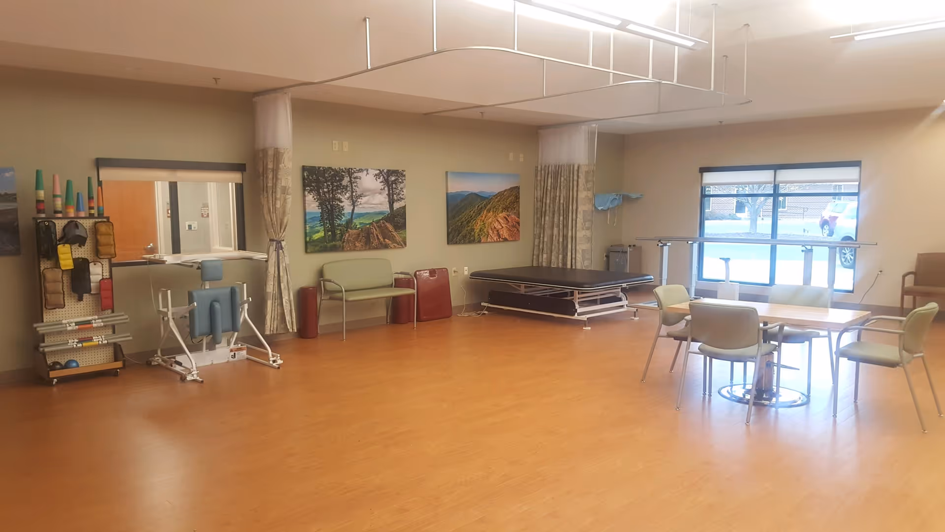 A spacious room with wooden flooring and beige walls, featuring physical therapy equipment including a padded exercise table, a blue therapy chair, and various exercise tools on a rack. There are two landscape paintings on the wall, a small green bench, a table with four chairs near a large window, and ceiling-mounted curtain tracks with curtains partially drawn.