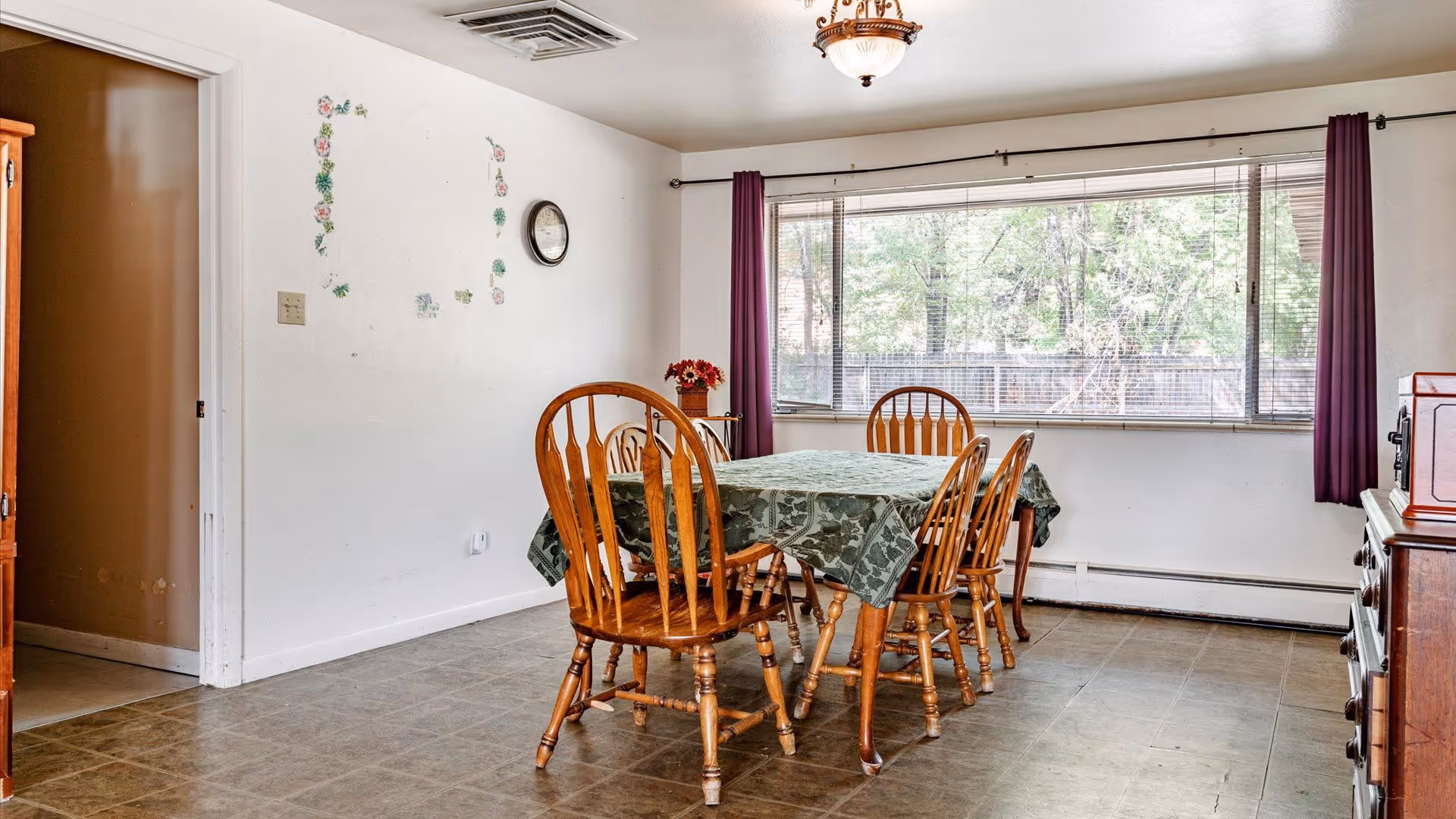 A dining room with a wooden table covered by a green patterned tablecloth and surrounded by six wooden chairs. There is a large window with purple curtains letting in natural light, a wall clock, and some floral wall decals on a white wall. The floor is tiled, and there is a wooden cabinet on the right side of the image.