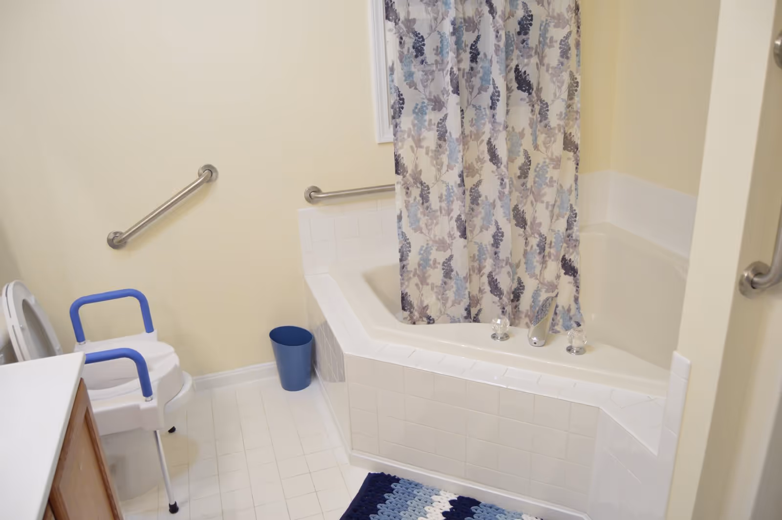 A bathroom with a white tiled bathtub featuring a floral shower curtain, a toilet with a blue-handled safety frame, grab bars on the walls, a blue trash bin, and a blue and white crocheted rug on the floor.