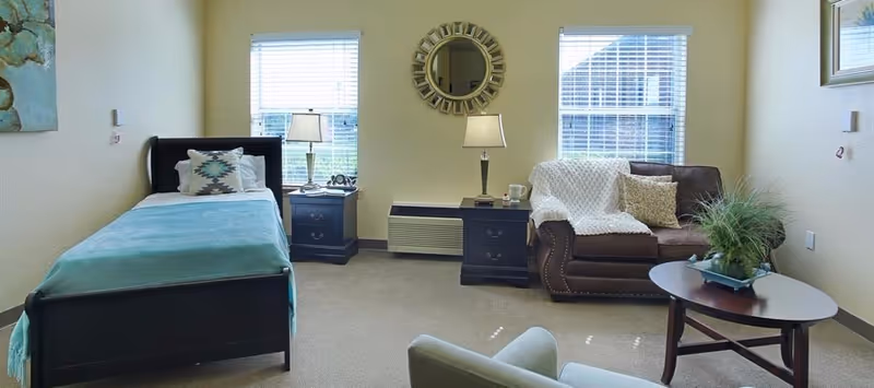 A cozy senior living room with a single bed on the left side, a nightstand with a lamp between two windows, a brown loveseat with pillows and a white throw blanket, a round coffee table with a plant, and a light green armchair in the foreground. A decorative round mirror hangs on the wall between the windows.