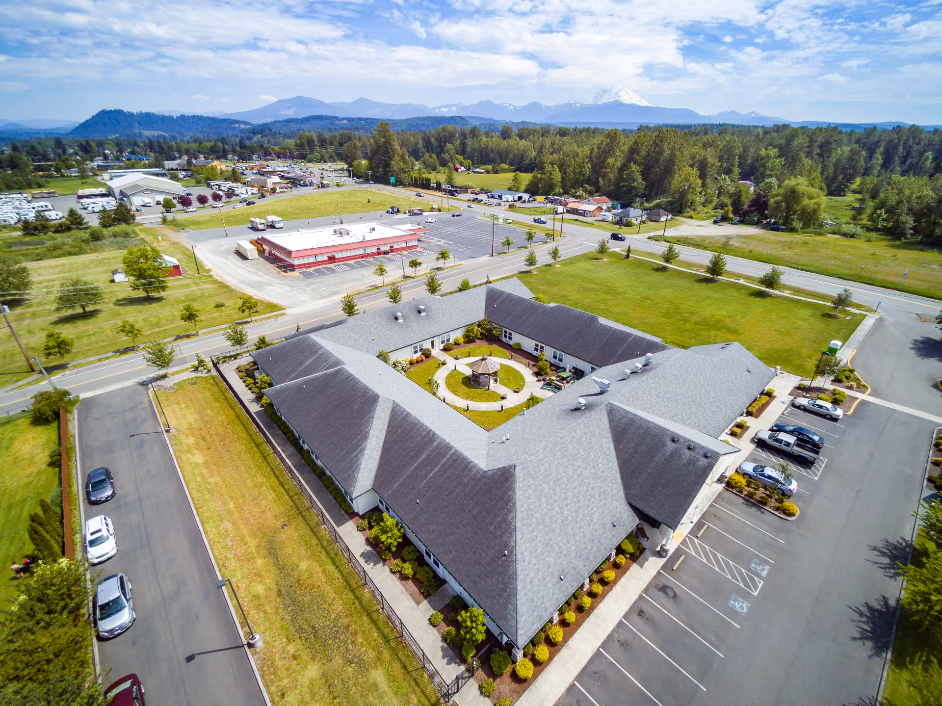 Aerial view of a single-story assisted living facility arranged around a central courtyard with parking lots, lawns, nearby roads and distant mountains.