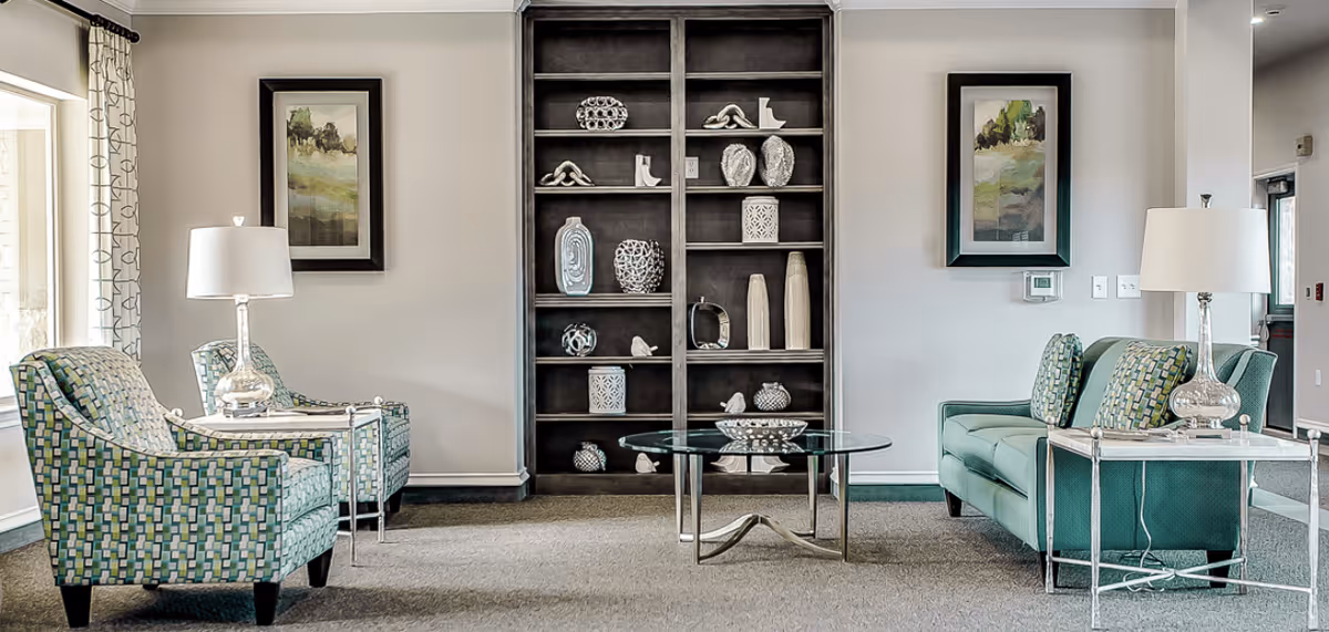 A cozy senior living common area with two patterned armchairs and a green sofa arranged around a glass coffee table. There are two side tables with lamps on either side of the seating. Behind the seating is a tall dark wooden bookshelf with decorative items and two framed landscape paintings on the wall.