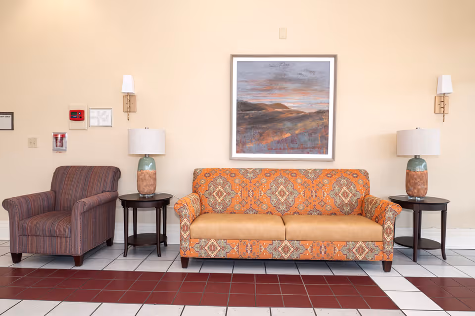 A seating area with a patterned orange and tan sofa and a striped armchair. Each side of the sofa has a dark wooden side table with a lamp featuring a green and brown base and a white lampshade. Above the sofa is a framed landscape painting with muted colors. The floor is tiled with white and red tiles, and the walls are painted beige.