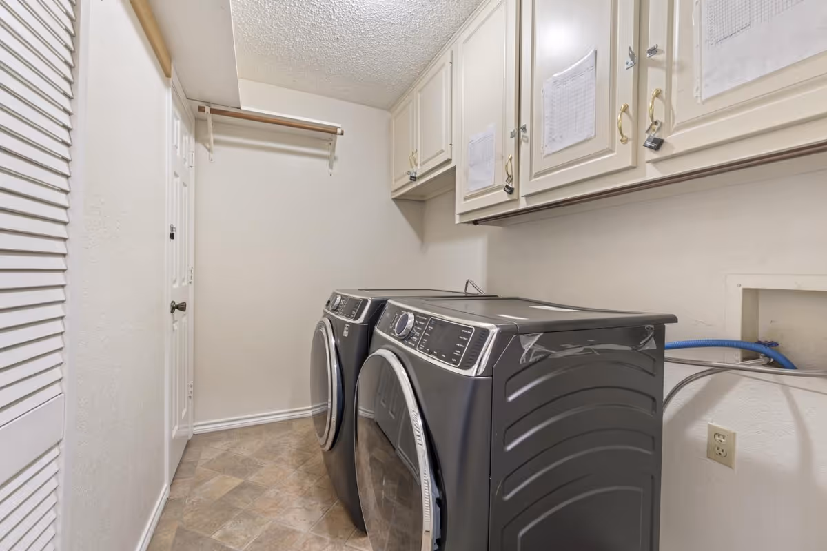 Laundry room with a modern front-loading washer and dryer set in a narrow space with beige tiled floor, white walls, white cabinets above the machines, and a wooden hanging rod on the wall.