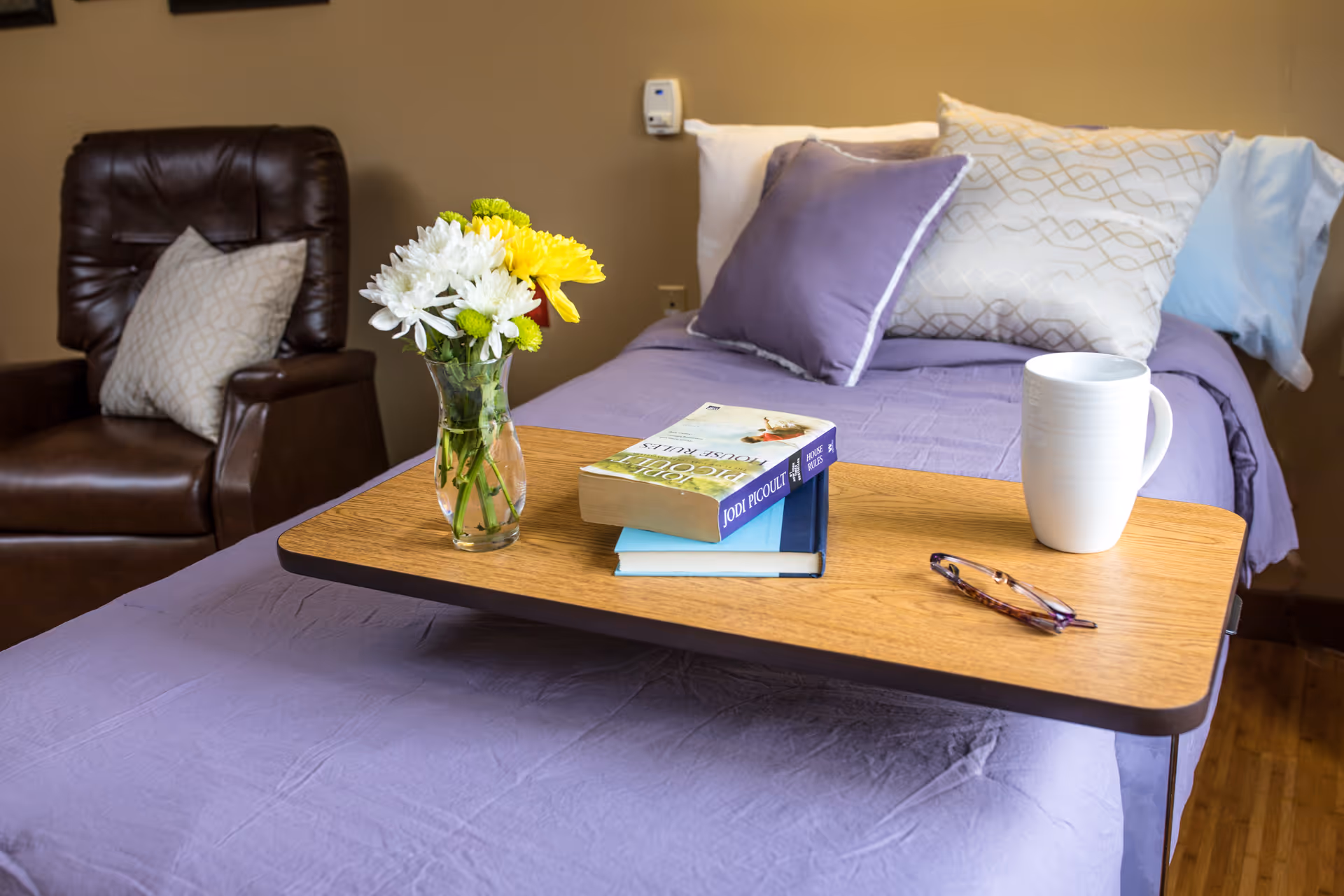 A cozy bedroom setup in an assisted living facility featuring a bed with purple bedding and multiple pillows, a wooden overbed table holding a vase of fresh flowers, two books, a pair of eyeglasses, and a white mug. A brown leather armchair with a patterned pillow is positioned next to the bed against a beige wall.