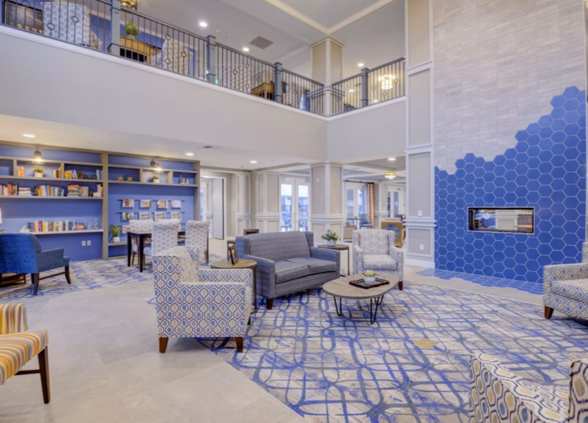 A spacious and well-lit senior living facility common area with a two-story ceiling. The room features a modern blue and gray color scheme, patterned armchairs, a gray loveseat, round wooden coffee tables, a blue hexagonal tiled fireplace, and built-in bookshelves with books and decorative items. There is a second-floor balcony overlooking the area.