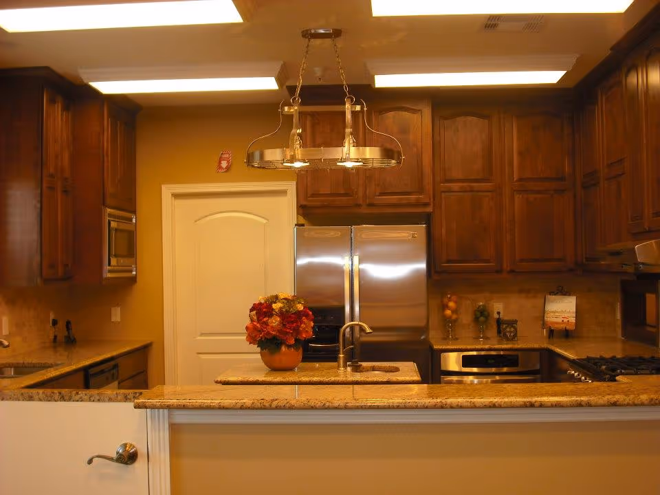 Interior view of a kitchen with wooden cabinets, a double-door stainless steel refrigerator, a built-in microwave, an oven, a gas stove, and a granite countertop island with a sink and a vase of flowers. The kitchen is well-lit with ceiling lights and a hanging light fixture above the island.