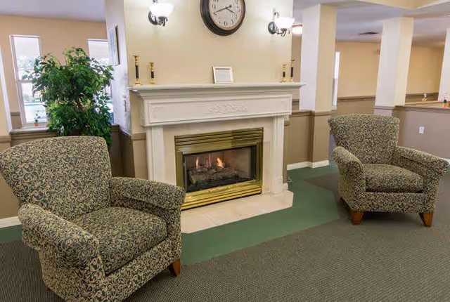 A cozy indoor seating area featuring two patterned armchairs positioned on either side of a white fireplace with a brass trim. Above the fireplace is a round clock and two wall-mounted light fixtures. A green plant is visible near a window in the background.