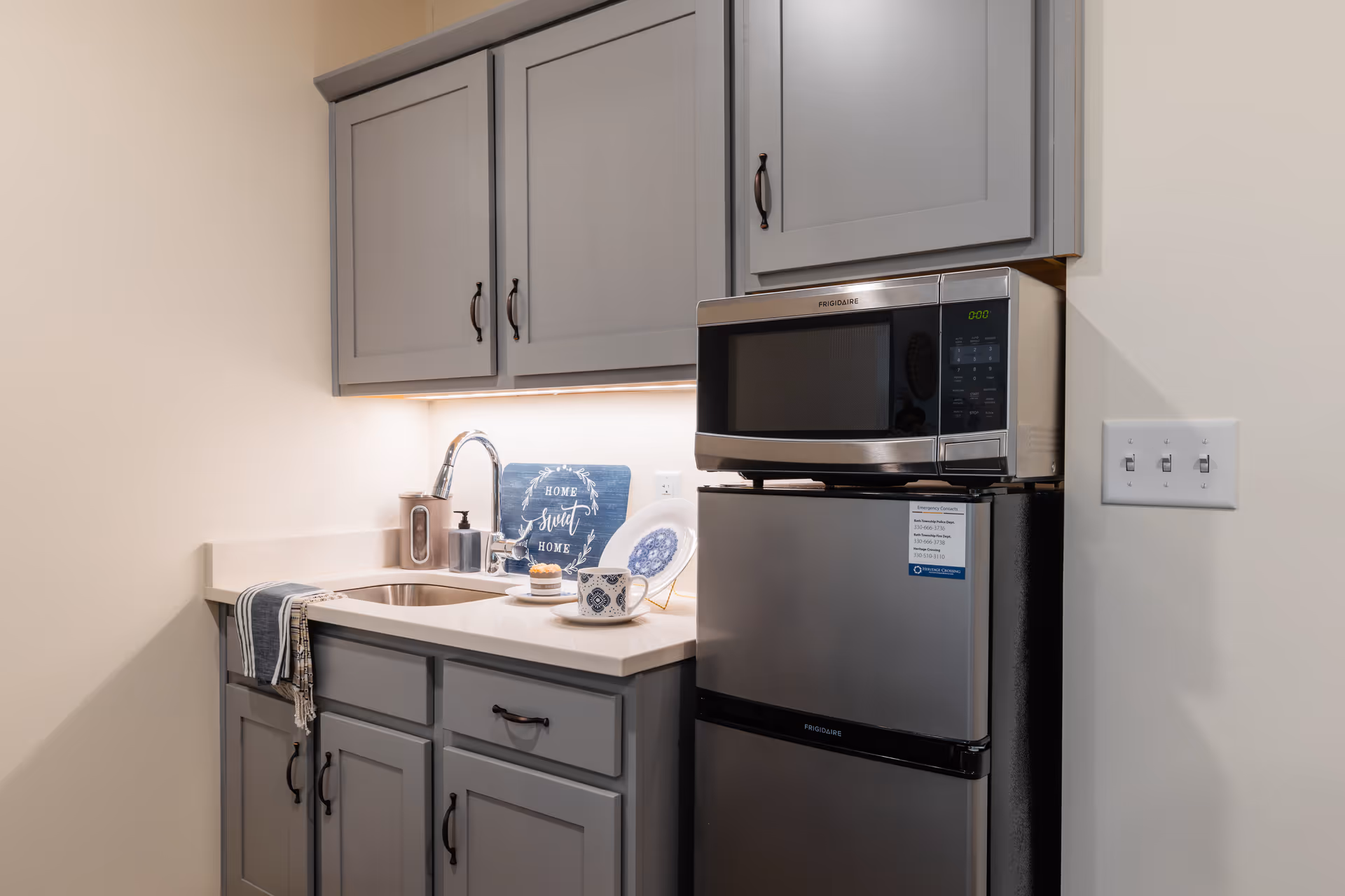 A small kitchen area with gray cabinets, a stainless steel sink, a silver microwave on top of a compact refrigerator, and a countertop with a decorative plate, a cup, and a small sign that reads 'Home Sweet Home'.