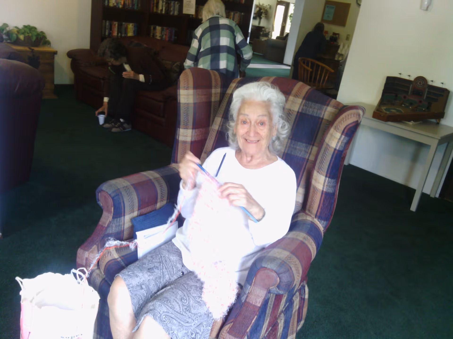 An elderly woman sitting in a plaid armchair knitting with pink yarn, smiling at the camera. In the background, two other elderly women are engaged in different activities in a cozy room with green carpet, brown leather sofas, bookshelves, and a table with a vintage radio.