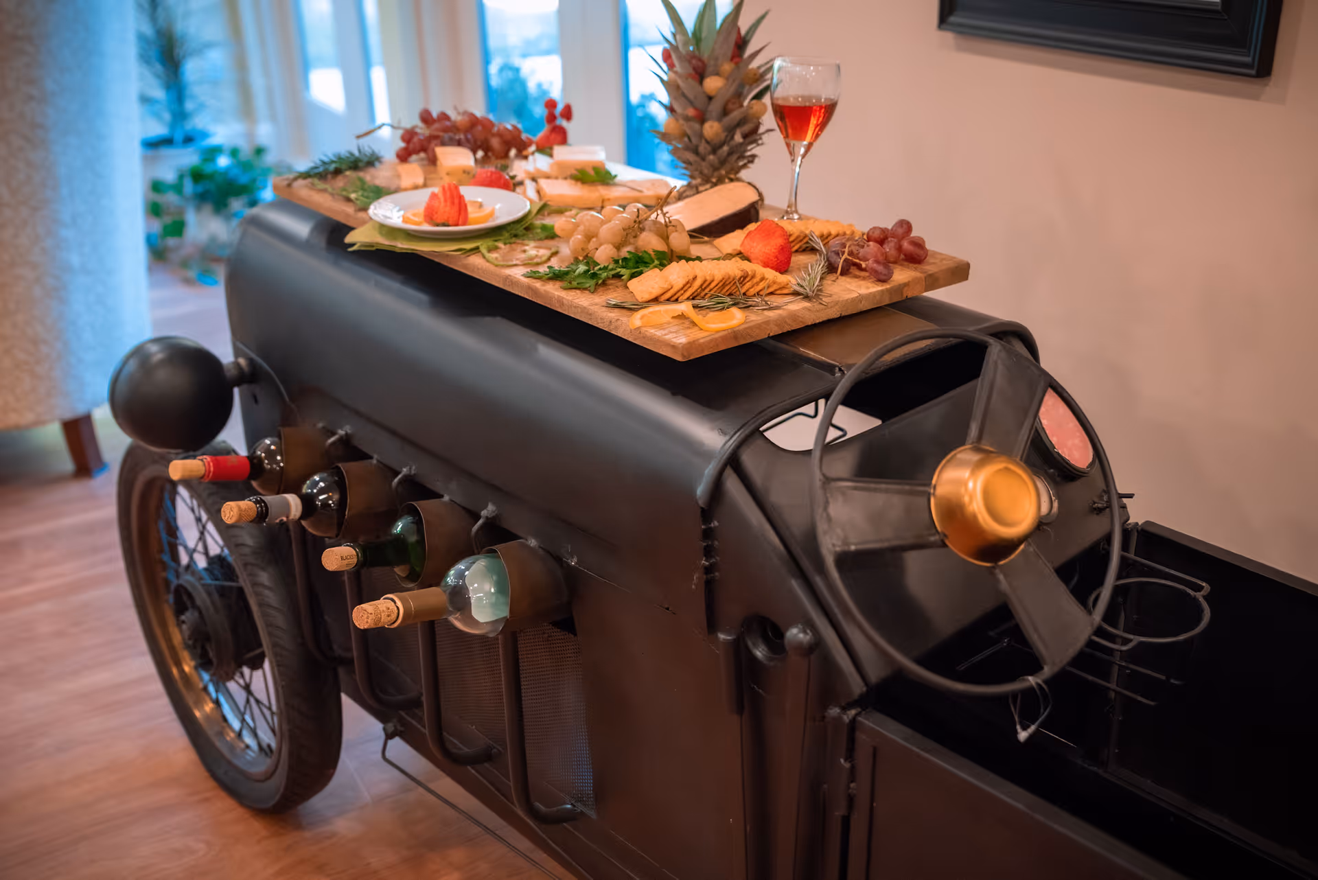 A vintage-style black metal cart designed to look like an old car, holding several bottles of wine on its side and topped with a wooden board displaying an assortment of cheeses, crackers, grapes, and a glass of red wine, set indoors on a wooden floor.