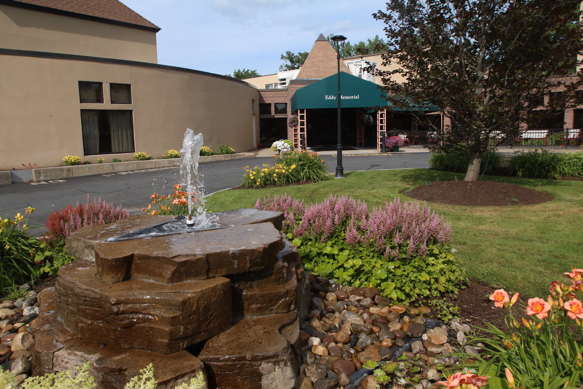 Outdoor view of the Terrace At Eddy Memorial facility featuring a stone water fountain surrounded by colorful flowers and greenery, with the building entrance visible in the background under a green awning.