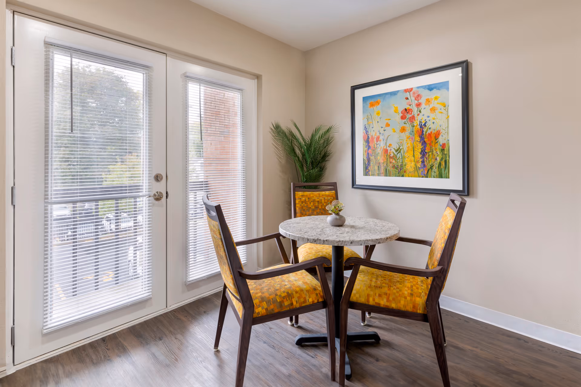 A small seating area with a round table and three wooden chairs with yellow patterned cushions. Behind the table is a potted plant and a colorful framed painting of flowers on the wall. To the left, there are glass double doors with blinds, letting in natural light.