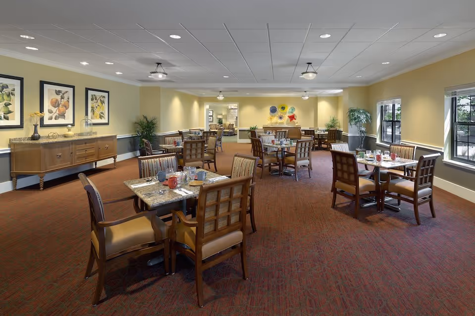 A spacious dining room with multiple tables and chairs arranged neatly. Each table is set with cups, glasses, and utensils. The room has warm yellow walls, large windows letting in natural light, framed fruit artwork on one wall, and decorative plants placed around the room. The carpet is a deep red color, and the ceiling has recessed lighting and hanging light fixtures.
