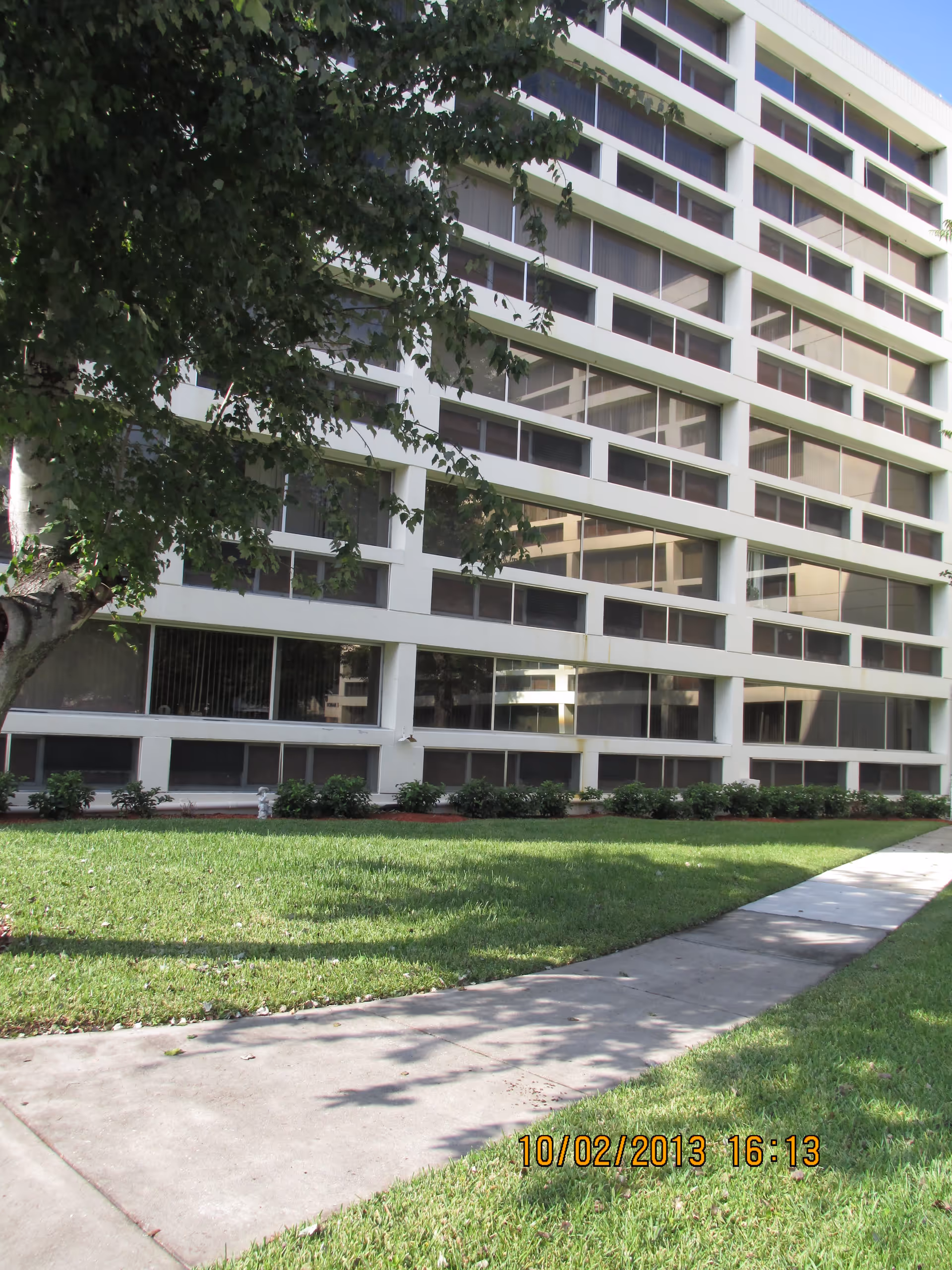 Exterior view of a multi-story building with large windows and white framing. In front of the building is a well-maintained lawn with a concrete walkway and a tree providing some shade.