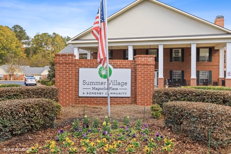 Entrance sign for Summer Village Magnolia Place, a Somerby community, mounted on a brick wall with an American flag in front and a flower bed with yellow and purple flowers. Behind the sign is a large building with white columns and a porch.