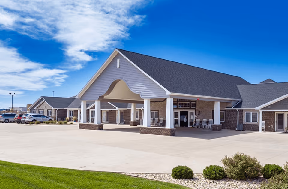 Exterior view of The Villas of Holly Brook Reflections Memory Care - Morton building with a large covered entrance, several windows, and a parking area with cars. The sky is clear with some clouds, and there is landscaping with green grass and bushes in the foreground.