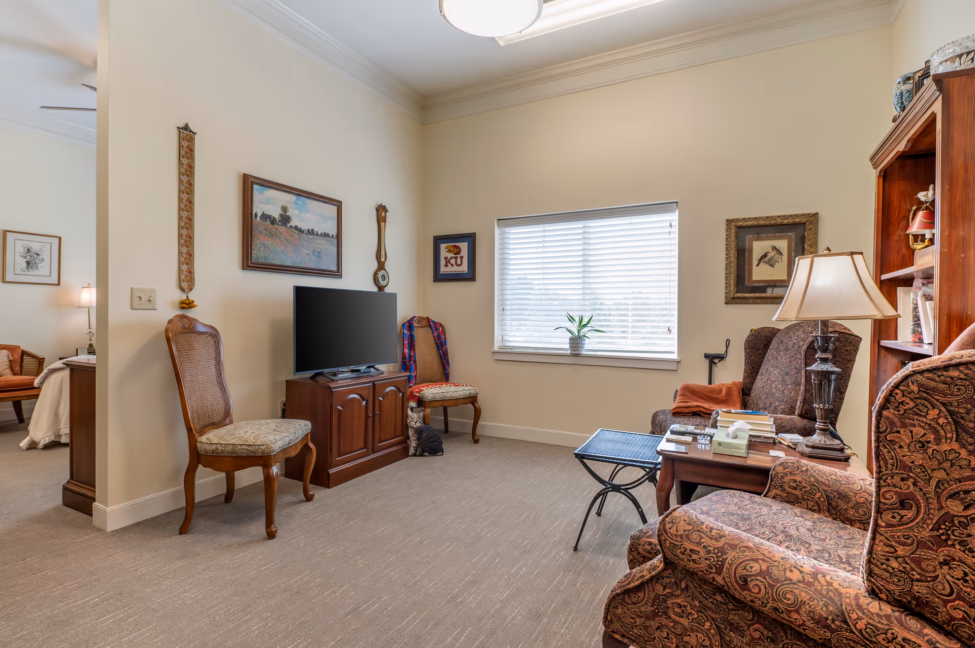 A cozy living room in a senior living facility with two patterned armchairs, a wooden side table with a lamp and books, a small black metal table, two wooden chairs with cushions, a flat-screen TV on a wooden cabinet, and a window with white blinds. The walls are decorated with framed pictures and a wall clock. A cat sits on the floor near the TV cabinet. An adjacent bedroom is partially visible through an open doorway.