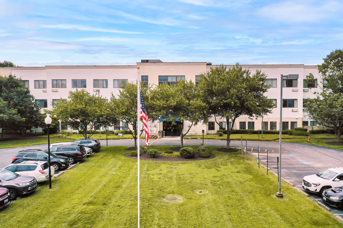 Front exterior view of a three-story senior living facility building with multiple windows, a central entrance, an American flag on a flagpole, green lawn, trees, and parked cars on either side of the driveway.
