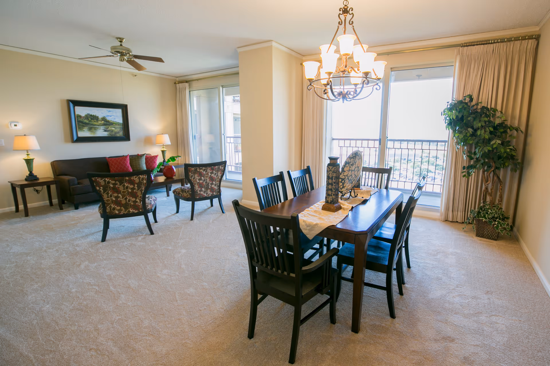 Bright open living and dining area with a wooden dining table and chandelier, seating area and sliding glass doors to a balcony.