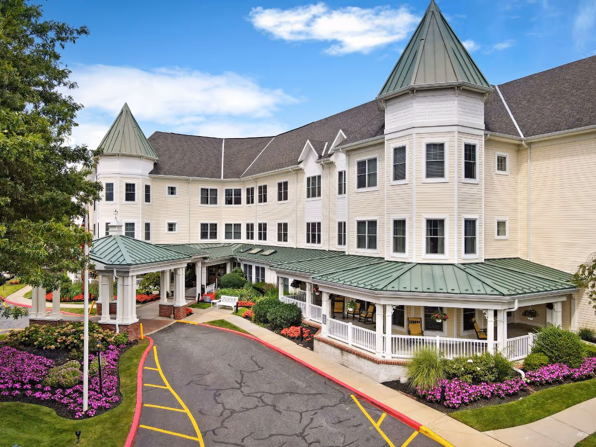 Front entrance of a large multi-story senior living building with turrets, a covered porte-cochere, and landscaped flower beds.
