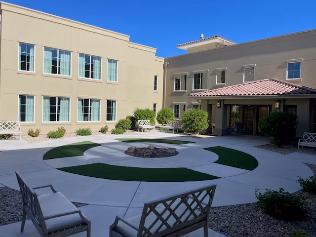Outdoor courtyard area of a senior living facility with circular concrete pathways and green artificial turf sections surrounding a central fire pit. The courtyard is enclosed by a two-story beige building with multiple windows and a covered entrance with a tiled roof. Several benches are placed around the courtyard, and there are small bushes and landscaping along the building walls.