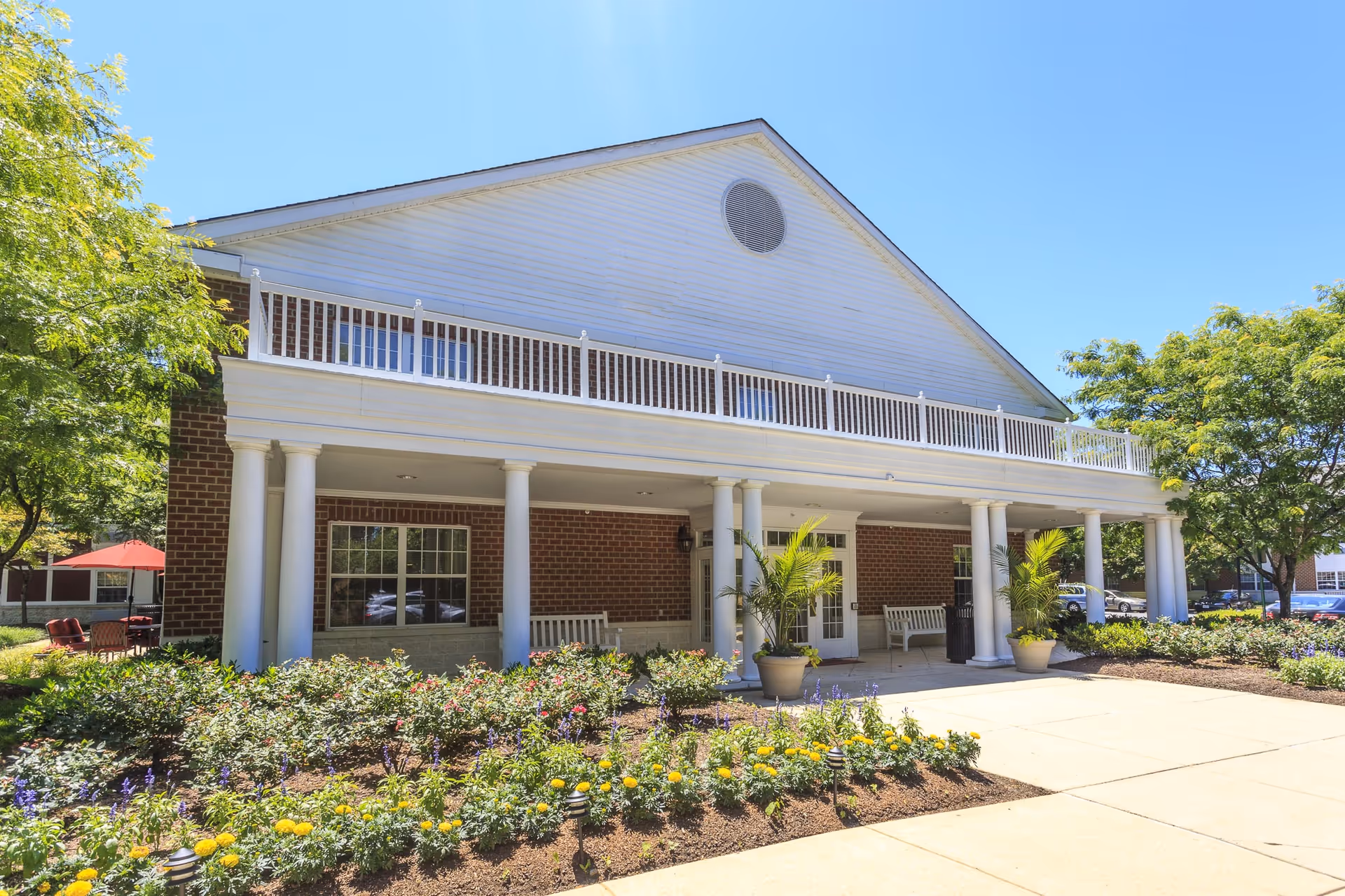 Two-story brick building with white columns and a covered porch entrance, surrounded by landscaped flower beds and trees.