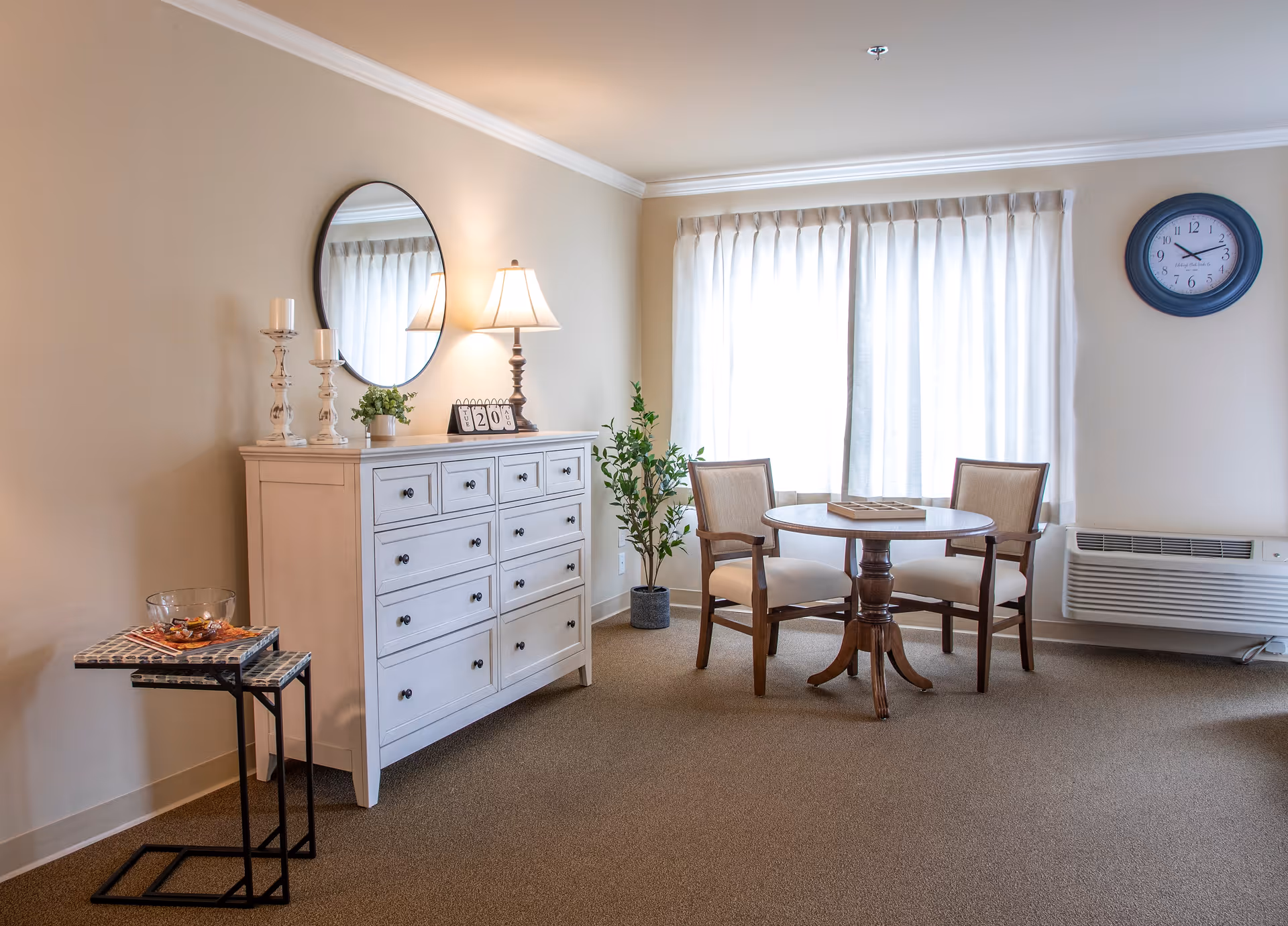A cozy interior room with a round wooden table and two upholstered chairs near a window with white curtains. A white dresser with multiple drawers is against the wall, topped with a round mirror, a table lamp, two candle holders, and a small plant. A small side table with a glass bowl and some items is next to the dresser. A wall clock and an air conditioning unit are visible on the right wall.