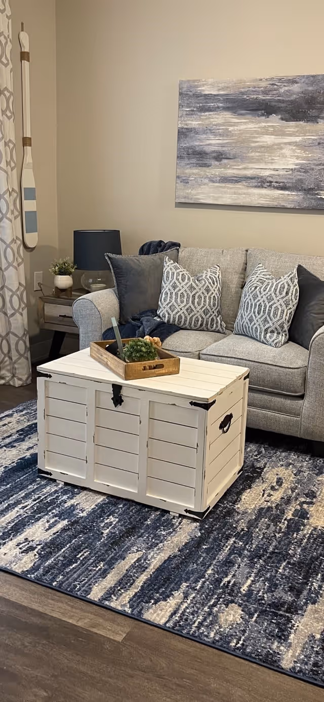 A cozy living room area featuring a light gray sofa with patterned and solid throw pillows, a white wooden chest used as a coffee table with a decorative tray on top, a blue and white patterned rug, a side table with a lamp and plant, and a large abstract painting on the wall.