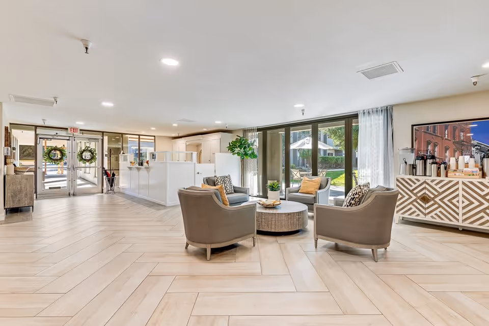 A bright and spacious senior living facility common area with a seating arrangement of four cushioned chairs around a round coffee table. Large windows with sheer curtains allow natural light to fill the room, and a sideboard with coffee and beverage supplies is visible on the right. The entrance doors with wreath decorations are seen in the background.