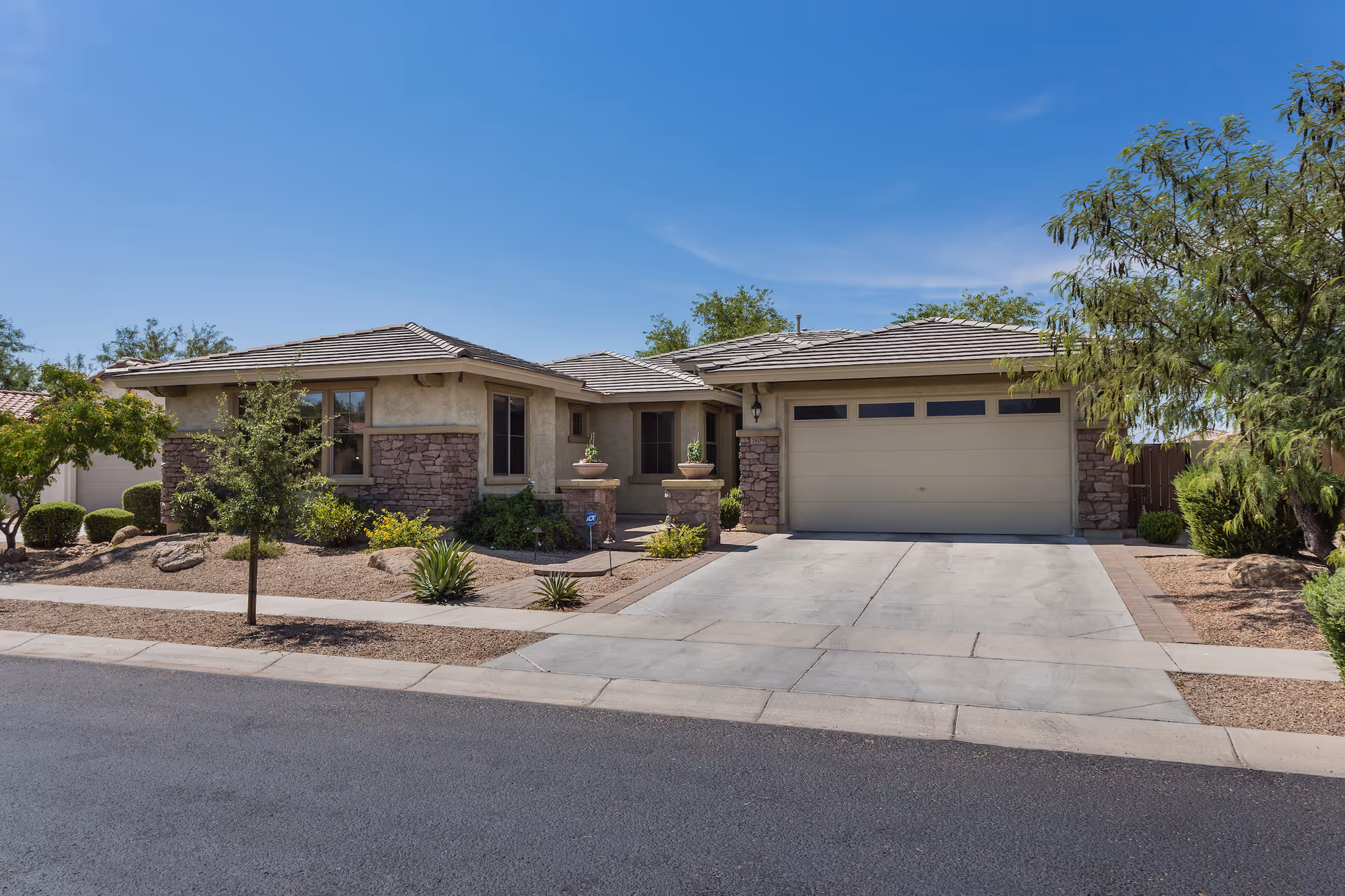 Exterior view of a single-story residential building with a two-car garage, stone and stucco facade, desert landscaping with small trees and shrubs, and a clear blue sky.