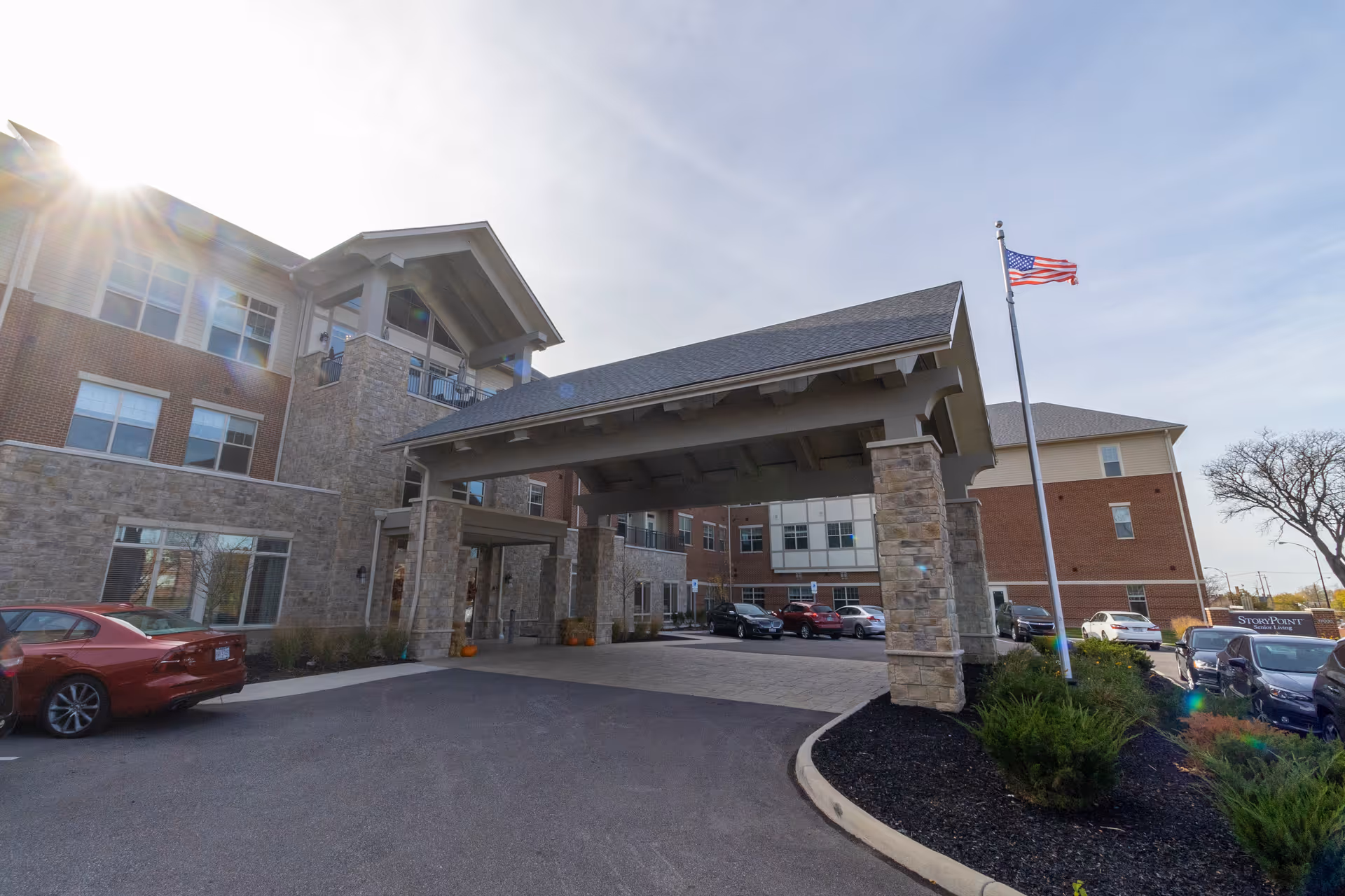 Front entrance of a multi-story brick and stone senior living building with a covered porte-cochere, American flag, and parked cars.