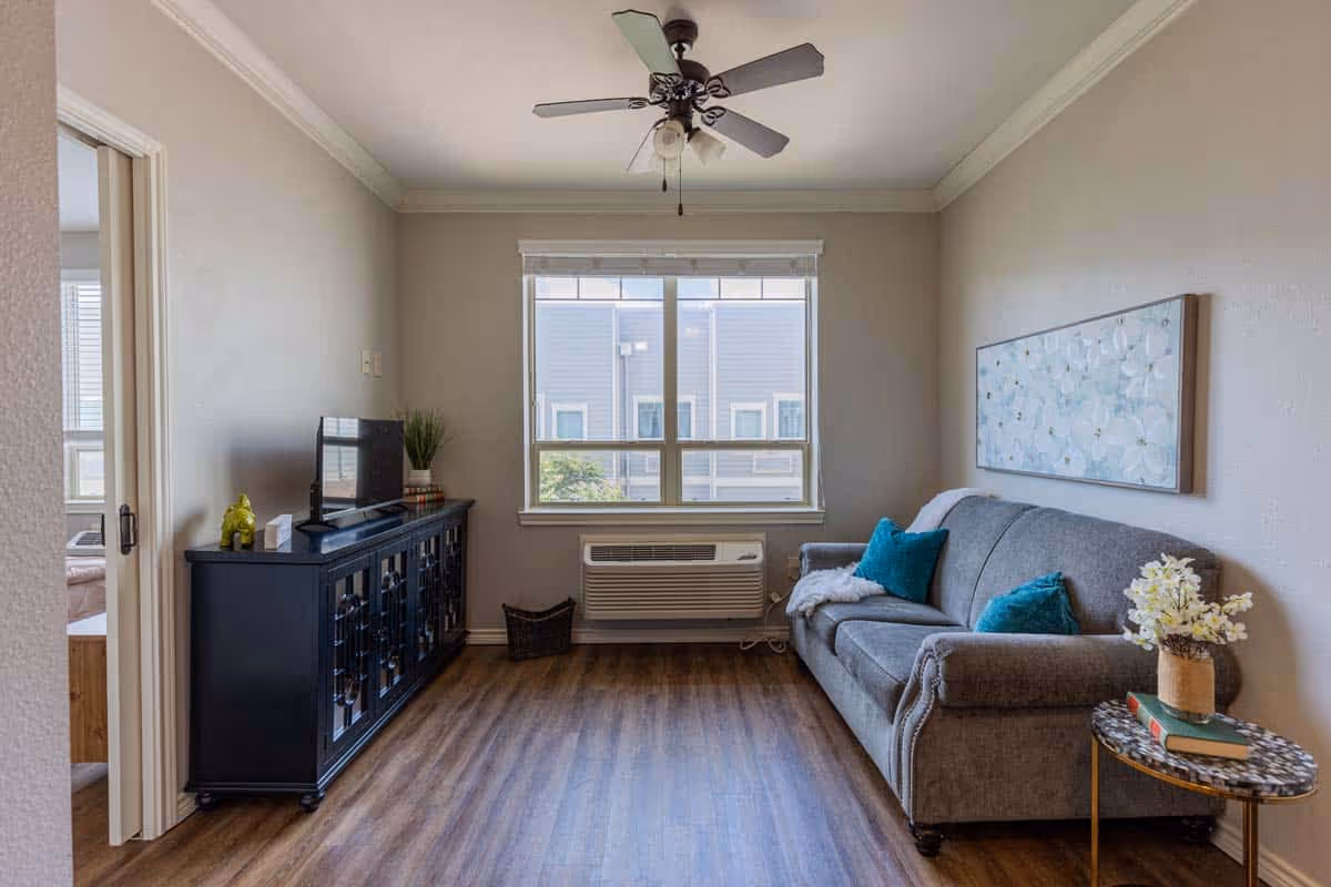 A cozy living room with a gray sofa adorned with teal pillows and a throw blanket, a small round side table with a vase of flowers and books, a black cabinet with a TV and decorative items, a window with blinds, and a ceiling fan above. The room has wood flooring and neutral-colored walls.