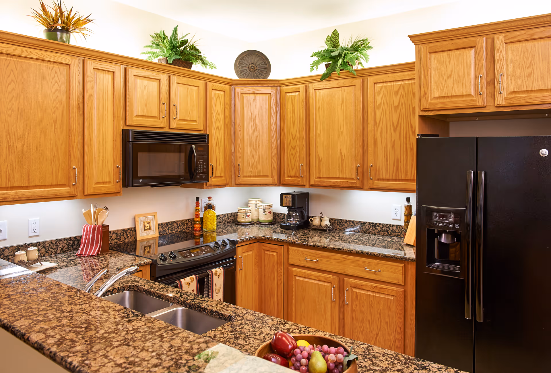 A kitchen with wooden cabinets, granite countertops, a black refrigerator with a water dispenser, a black microwave above a stove, a double sink, and various kitchen items including a coffee maker, jars, and a bowl of fruit on the counter.