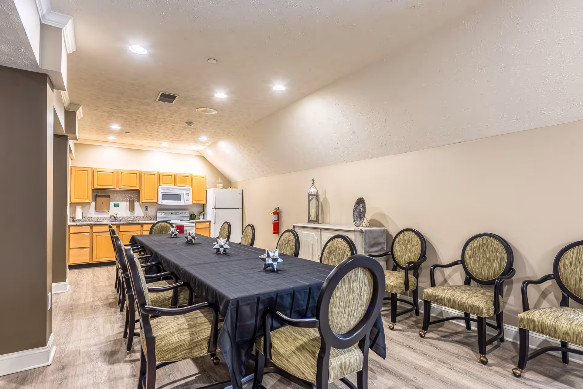 A dining area with a long rectangular table covered with a black tablecloth, surrounded by green cushioned chairs with black frames. In the background, there is a kitchen area with wooden cabinets, a white microwave, stove, and refrigerator. The room has light-colored walls and wood flooring, with ceiling lights illuminating the space.