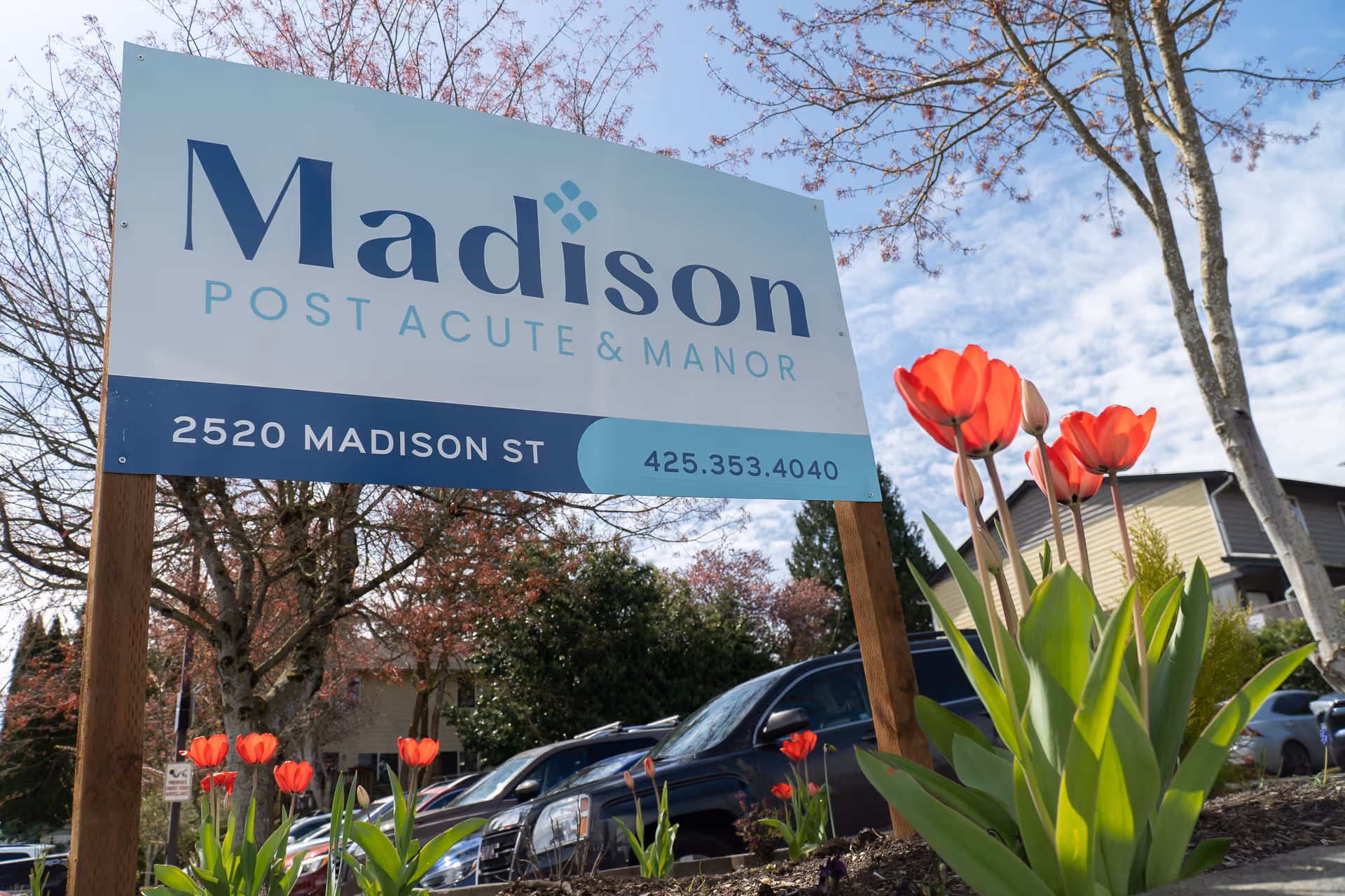 Outdoor view of a sign for Madison Post Acute & Manor located at 2520 Madison St with a phone number 425.353.4040. The sign is surrounded by blooming red tulips and trees with a partly cloudy sky in the background. Cars and a building are visible behind the sign.