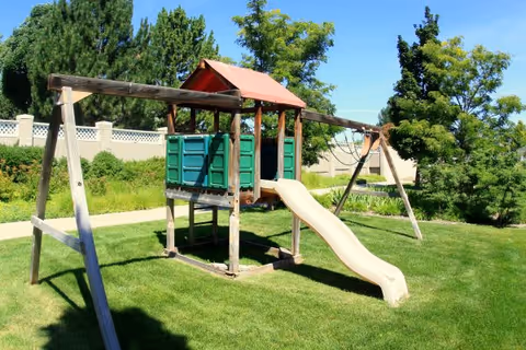 A wooden playground structure with a slide, swings, and a small covered platform situated on a grassy area with trees and a fence in the background under a clear blue sky.