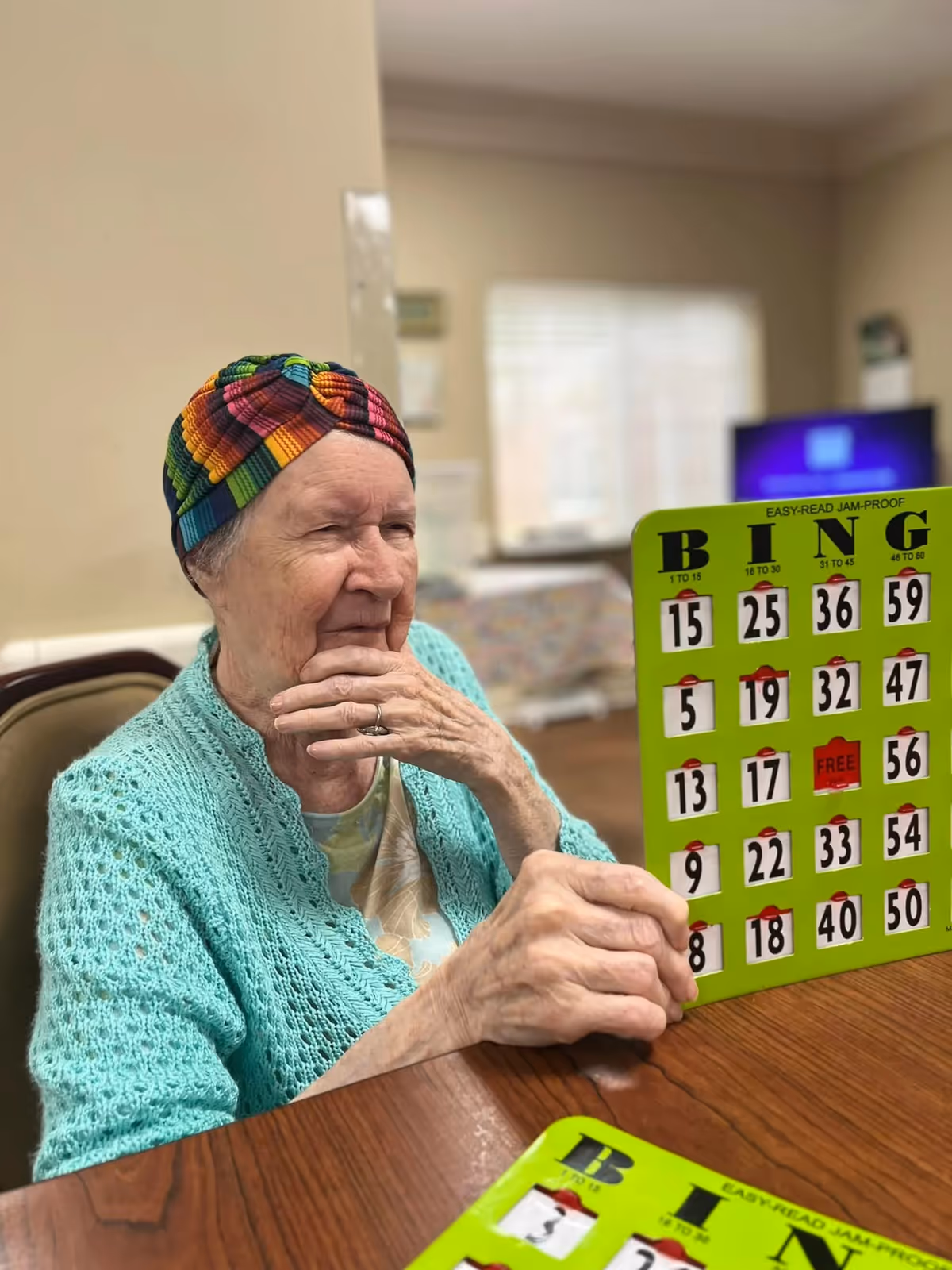 An elderly woman wearing a colorful headwrap and a light blue knitted cardigan is sitting at a wooden table playing bingo. She is holding a green bingo card and appears to be concentrating on the game. The background shows a softly lit room with a window and a television.