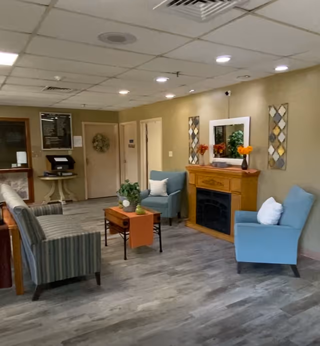 Communal lounge with armchairs, a striped sofa, coffee table, decorative fireplace and wall art under recessed ceiling lights.