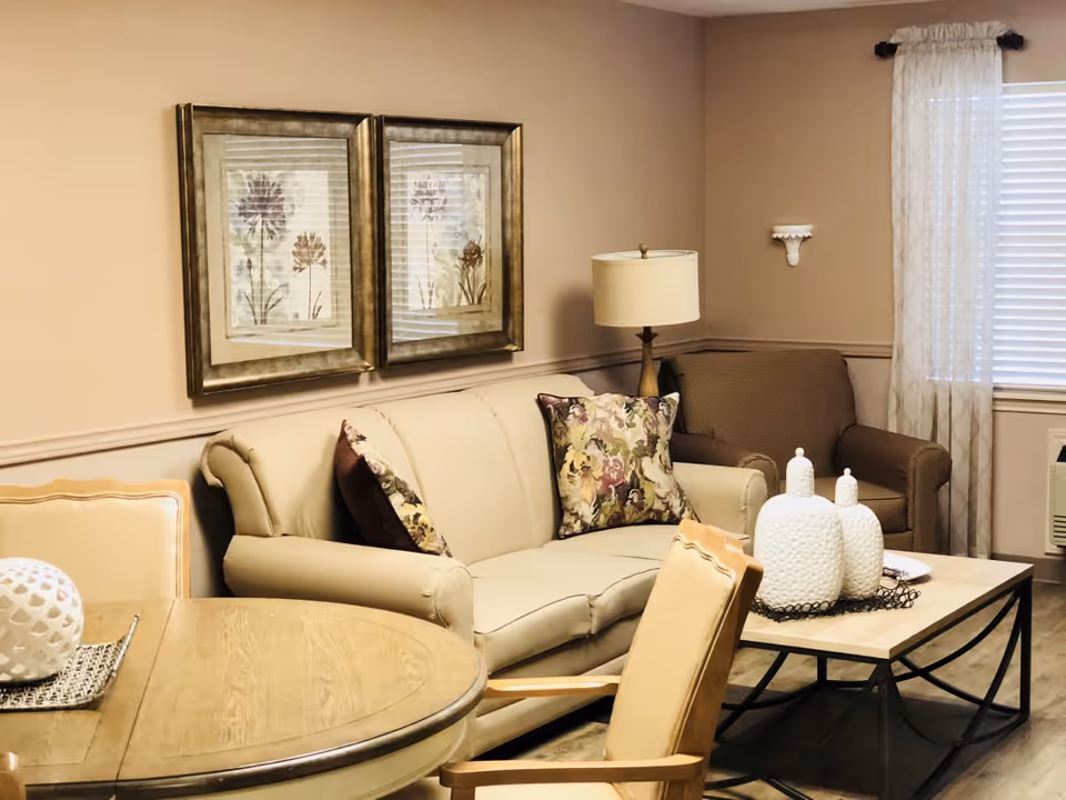 A cozy living room area with a beige sofa adorned with floral patterned pillows, a brown armchair, a wooden coffee table with decorative white ceramic jars, a round wooden dining table with chairs, two framed floral artworks on the wall, a standing lamp, and a window with sheer curtains.
